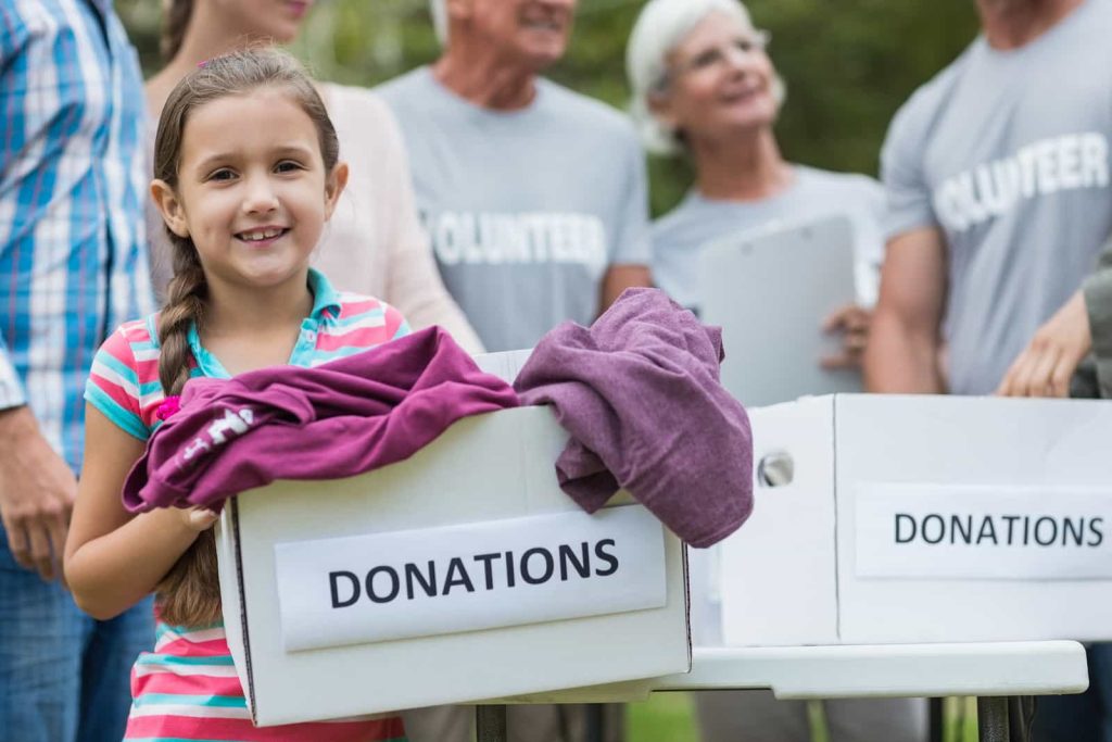 A smiling young girl holding a donation box with clothes stands in the forefront. Behind her, a group of people wearing "Volunteer" t-shirts are gathered, some holding clipboards. Two white boxes labeled "Donations" are on the table.