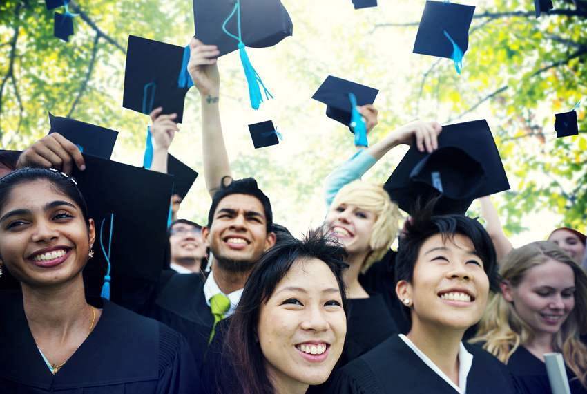 A joyful group of graduates in caps and gowns celebrate by throwing their mortarboards into the air. They are standing outdoors on a sunny day, smiling and looking upwards, with trees and sunlight filtering through the leaves in the background.