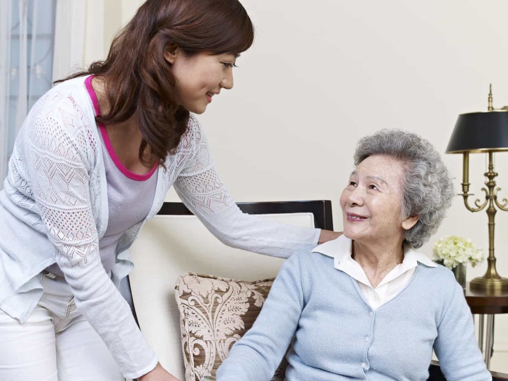 A younger woman stands beside an elderly woman who is seated. Both women are smiling at each other warmly. The elderly woman has gray hair and wears a light blue sweater, while the younger woman has long brown hair and wears a white cardigan over a pink top.