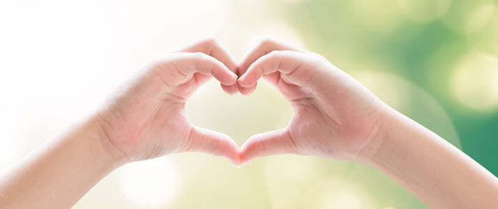 Two hands form a heart shape against a softly blurred green and white background.