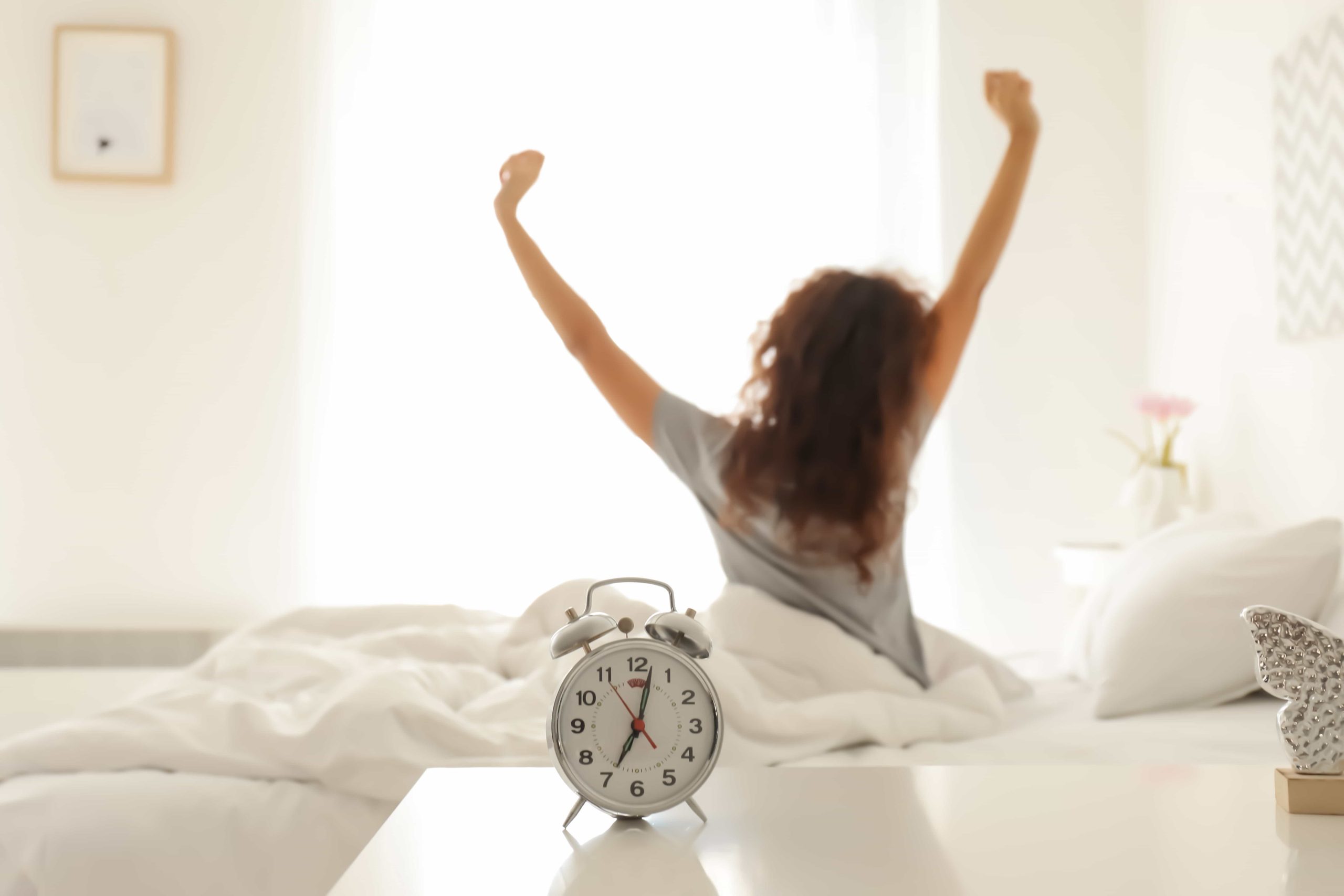 A woman sits on a bed, stretching her arms upwards with a feeling of refreshment. a classic alarm clock shows the time in a warmly lit bedroom.